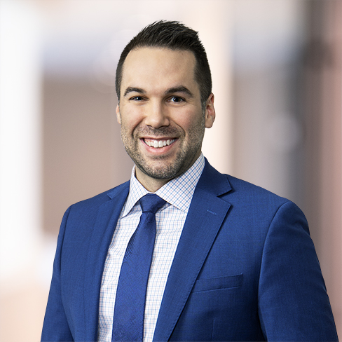 A man wearing a blue suit, light checkered shirt, and blue tie is smiling at the camera in a softly blurred indoor setting, reflecting the professional atmosphere of a corporate law office.