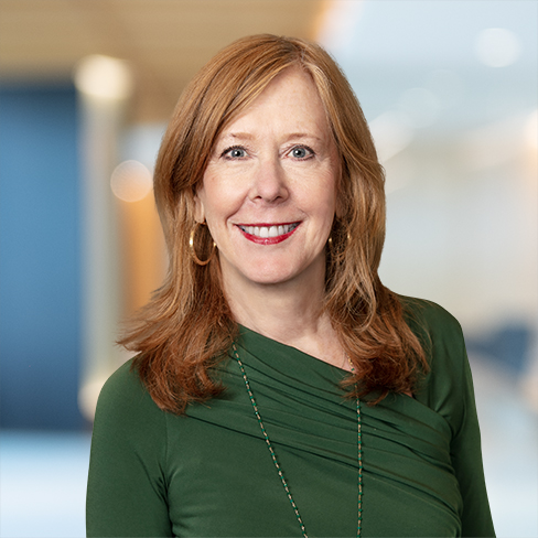 Woman with long red hair wearing a green top stands indoors, smiling at the camera. The blurred background suggests an office or professional setting, possibly related to chicago lawyers or litigation support services.