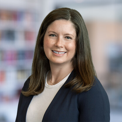 A woman with straight brown hair, wearing a navy blazer and light shirt, smiles at the camera. The softly blurred background hints at a corporate law office in Chicago, creating a professional atmosphere.