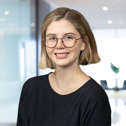 A young woman with short blonde hair and round glasses wearing a black sweater smiles in a modern corporate law office setting.