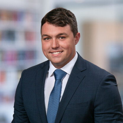 A man in a dark blue suit, white shirt, and blue tie is smiling in front of a blurred office background, reflecting the professionalism of a corporate law office.