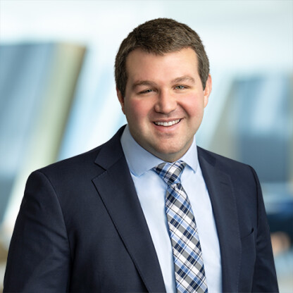 Man in a dark suit and plaid tie smiling at the camera, standing in a modern office setting with blurred background, representing experienced Chicago lawyers specializing in intellectual property law.