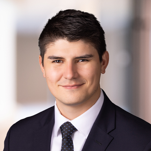 A man wearing a black suit, white shirt, and dotted tie, with short dark hair, poses for a professional headshot against a blurred background—ideal for representing law offices or corporate law office environments.