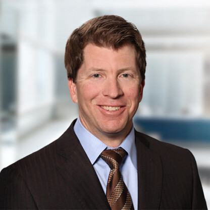 A man in a dark suit, light blue shirt, and patterned brown tie smiles in a bright, modern corporate law office setting.