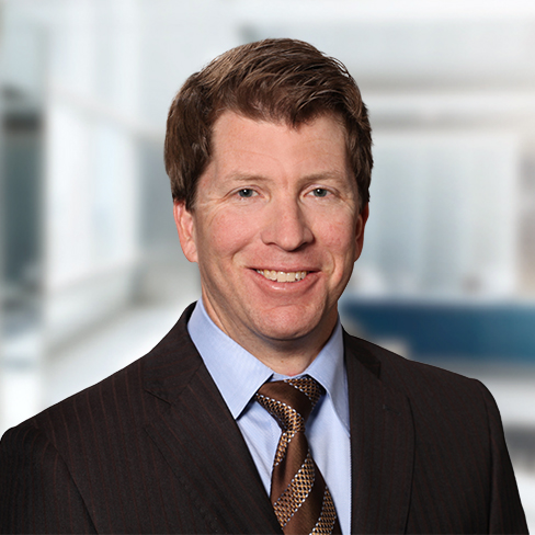 A man in a dark suit, light blue shirt, and patterned brown tie smiles in a bright, modern corporate law office setting.