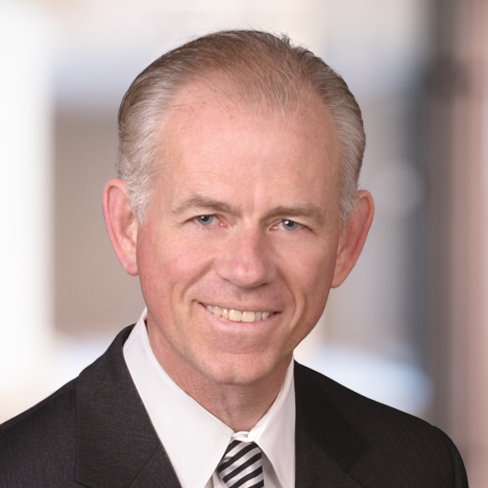 A middle-aged man with short gray hair, wearing a dark suit, white shirt, and striped tie, smiles at the camera against a blurred indoor background—representing experienced Chicago lawyers offering trusted litigation support.
