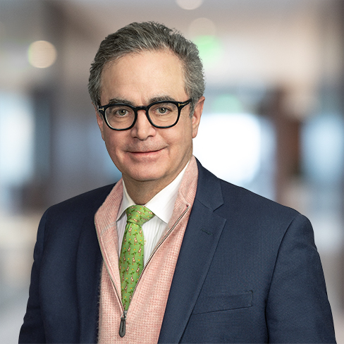 A man with gray hair and glasses, dressed in a navy suit, pink sweater, white shirt, and green tie, stands in a blurred indoor setting typical of Chicago lawyers or law offices.