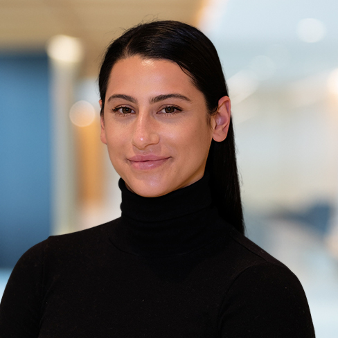 A woman with long dark hair and a black turtleneck top stands indoors, with the blurred backdrop of a corporate law office behind her.