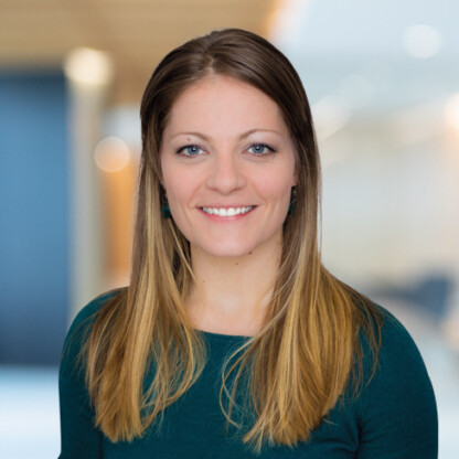 A woman with straight, light brown hair wearing a dark green top smiles at the camera in a softly blurred corporate law office setting.