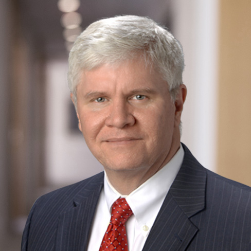 A middle-aged man with short gray hair wearing a dark pinstripe suit, white shirt, and red tie stands confidently in the blurred hallway of a corporate law office.