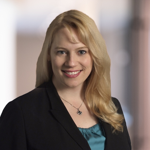 A woman with long blonde hair wearing a black blazer, teal blouse, and a necklace, smiling at the camera with a blurred indoor corporate law office background.
