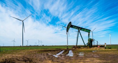 An oil pump jack operates in a muddy field with several wind turbines in the background, reminiscent of the dynamic energy clients handled by Chicago lawyers under a blue sky with scattered clouds.