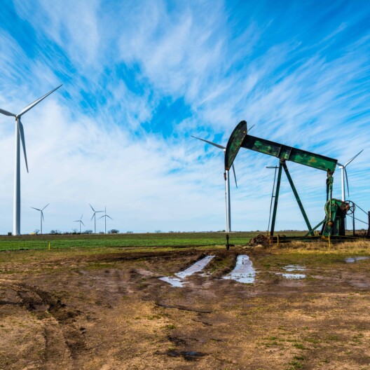 An oil pump jack operates in a muddy field with several wind turbines in the background, reminiscent of the dynamic energy clients handled by Chicago lawyers under a blue sky with scattered clouds.