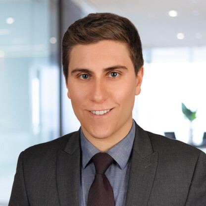 A young man in a gray suit and dark tie smiles at the camera in a modern corporate law office with glass walls and a plant in the background, reflecting expertise in litigation support.