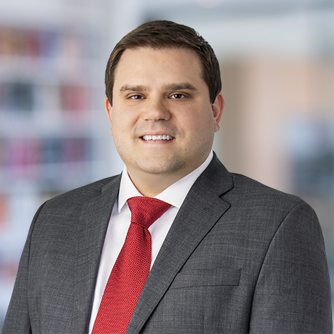 A man in a gray suit, white shirt, and red tie stands in an office setting with shelves blurred in the background, reflecting the professional atmosphere of law offices specializing in litigation support.