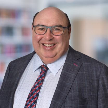 A middle-aged man with glasses, wearing a plaid suit jacket, light dress shirt, and patterned tie, smiles in a blurred office or library setting—reflecting the professionalism of Chicago lawyers specializing in litigation support.