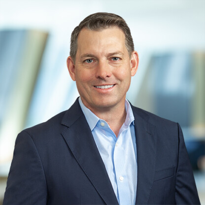 A man in a navy suit and light blue shirt smiles while standing in front of a blurred corporate law office background.