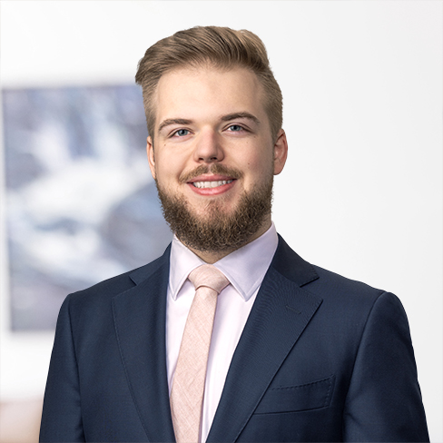 A young man with light hair and a beard is wearing a navy suit, light pink shirt, and matching tie, standing in front of a blurred office background typical of Chicago lawyers or law offices.