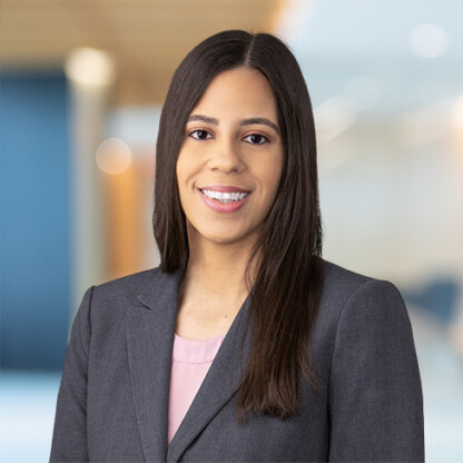 A woman with long straight hair, wearing a gray blazer and pink top, smiles at the camera in a modern corporate law office, reflecting the professional atmosphere of Chicago lawyers.