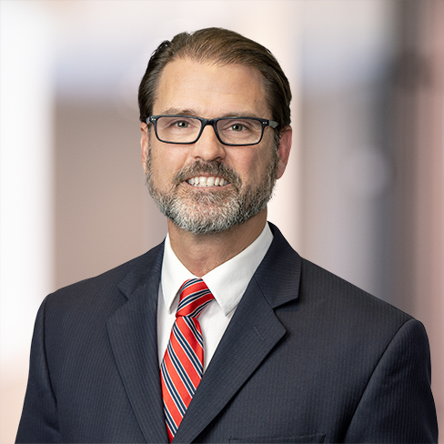 A man with glasses, a beard, and brown hair wearing a dark suit, white shirt, and red striped tie poses in front of a blurred office background, representing chicago lawyers specializing in litigation support.