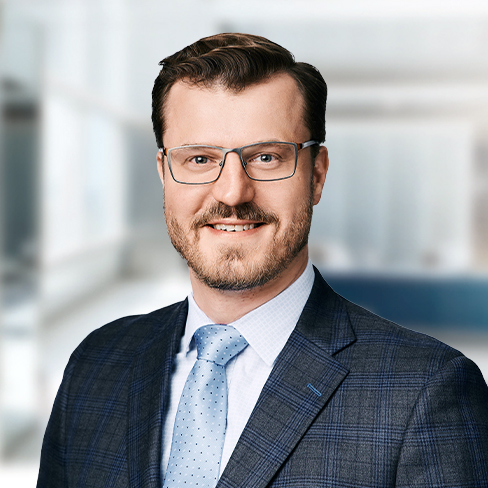 A man with short brown hair, glasses, and a trimmed beard, dressed in a blue suit, light blue shirt, and polka-dot tie stands in a bright office setting, typical of professional law offices offering litigation support.