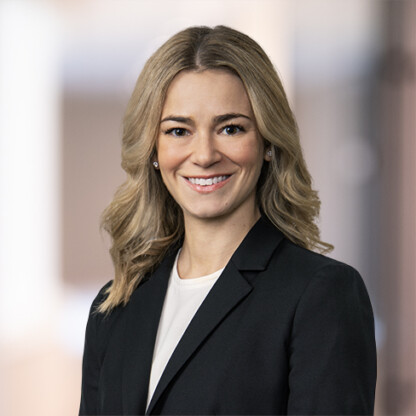 Woman with blonde hair wearing a black blazer and white top, smiling, posed in front of a blurred office background—an approachable professional at a leading corporate law office.