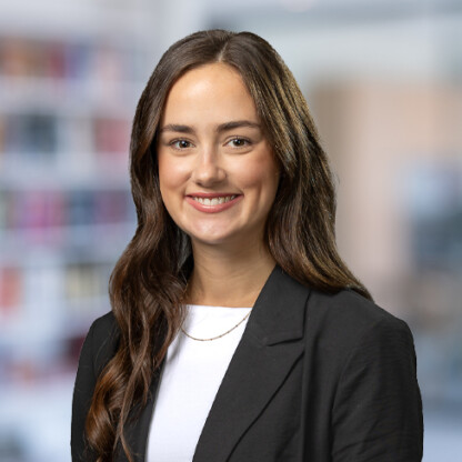 Young woman with long brown hair wearing a black blazer and white top, smiling at the camera in a blurred corporate law office background.