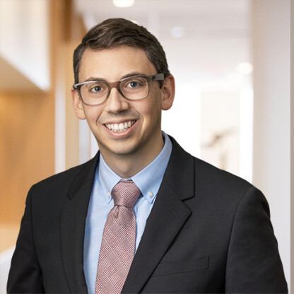A man wearing glasses, a dark suit, a light blue shirt, and a patterned tie stands in the brightly lit hallway of a corporate law office, smiling at the camera.