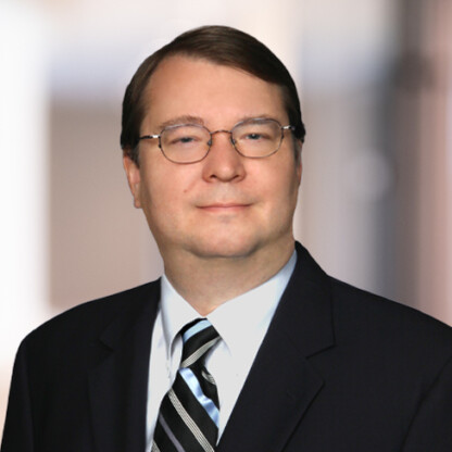 A man wearing glasses, a dark suit, white shirt, and striped tie stands in front of a blurred indoor background, embodying the professionalism of Chicago lawyers specializing in intellectual property law.