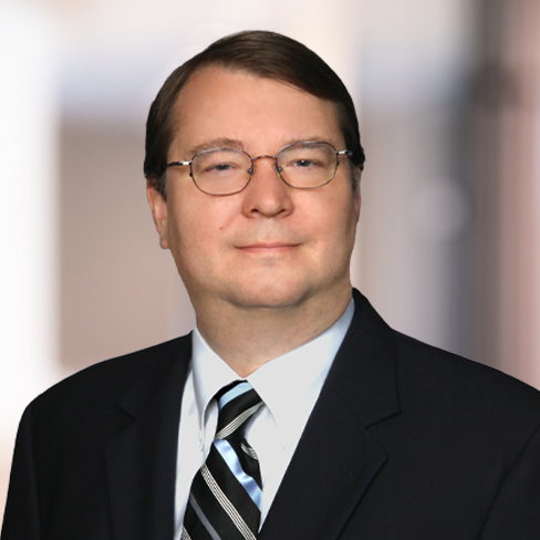 A man wearing glasses, a dark suit, white shirt, and striped tie stands in front of a blurred indoor background, embodying the professionalism of Chicago lawyers specializing in intellectual property law.