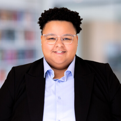 A person wearing glasses, a light blue dress shirt, and a black blazer stands indoors with a blurred background of shelves, evoking the professional atmosphere often seen in law offices or among lawyers in Chicago.