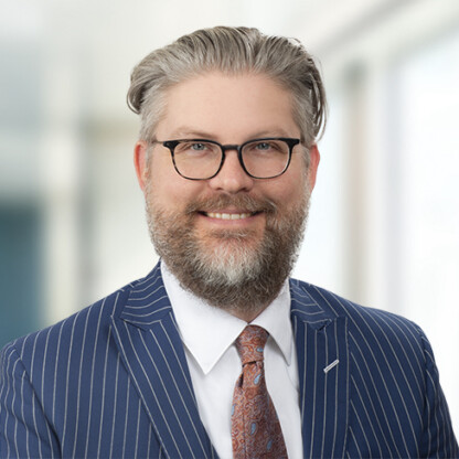 A man with glasses and a gray beard, dressed in a pinstripe suit, white shirt, and patterned tie, stands in front of a blurred office background—reflecting the professionalism found in top Chicago lawyers' law offices.
