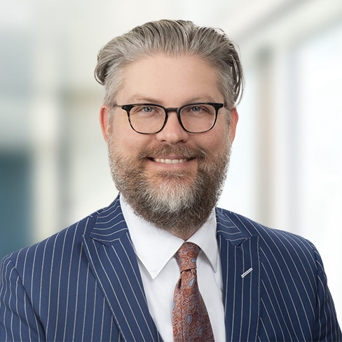 A man with glasses and a gray beard, dressed in a pinstripe suit, white shirt, and patterned tie, stands in front of a blurred office background—reflecting the professionalism found in top Chicago lawyers' law offices.