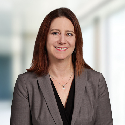 A woman with straight brown hair, wearing a gray blazer over a black top, smiles in front of a blurred indoor background at a corporate law office.
