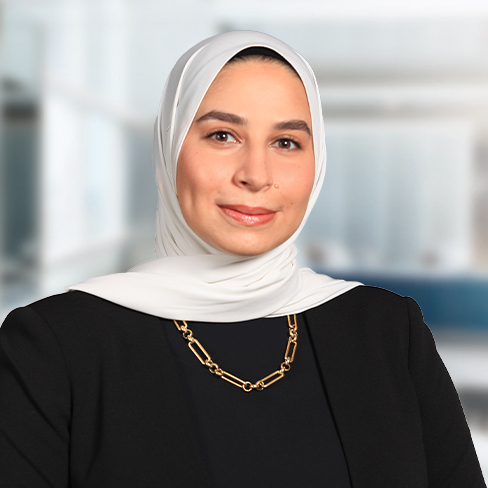 A woman in a white hijab and black blazer is smiling slightly, wearing a gold chain necklace, with the blurred backdrop of a corporate law office.