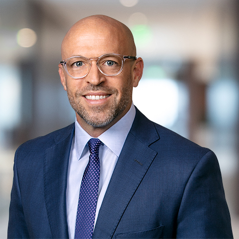 A man wearing a blue suit, light blue shirt, and glasses is smiling at the camera in a blurred corporate law office setting.