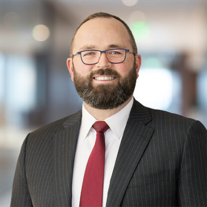 A man wearing a dark pinstripe suit, white shirt, and red tie stands indoors in a corporate law office, smiling at the camera.
