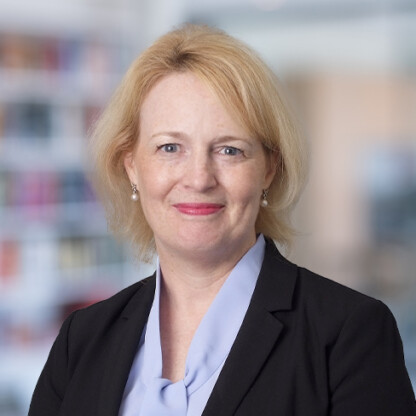 A woman with short blonde hair, wearing a black blazer and light blue blouse, smiles at the camera. A blurred corporate law office setting is in the background.