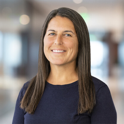 A woman with straight, long brown hair wearing a dark navy sweater stands indoors, smiling, with a blurred law offices background.
