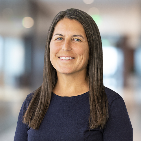 A woman with straight, long brown hair wearing a dark navy sweater stands indoors, smiling, with a blurred law offices background.