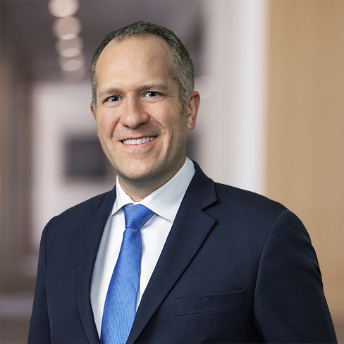 A man in a dark suit and blue tie stands in front of a blurred indoor background, smiling at the camera—capturing the professional atmosphere of a corporate law office.