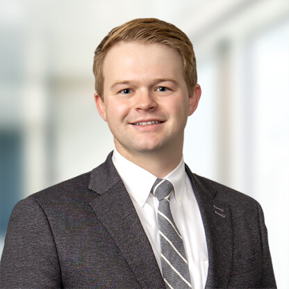 A young man in a dark suit, white shirt, and striped tie stands indoors with a blurred office background, reflecting the professional atmosphere of Chicago law offices. He is smiling and facing the camera.