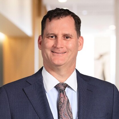 A man in a navy suit, white shirt, and patterned tie stands indoors with a neutral expression, set against the blurred backdrop of a corporate law office.