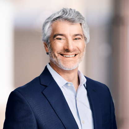Smiling man with gray hair and beard, wearing a navy blazer and light blue shirt, stands in front of a blurred indoor background—an approachable face often seen in top Chicago lawyers’ law offices offering trusted litigation support.