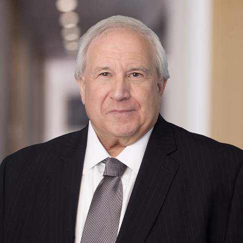 An older man with gray hair, wearing a dark pinstripe suit, white shirt, and gray tie, stands in a blurred hallway background of a corporate law office.