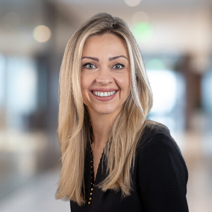 Woman with long blonde hair wearing a black top, smiling, standing in a modern, blurred indoor office setting—perfect for law offices or litigation support environments.