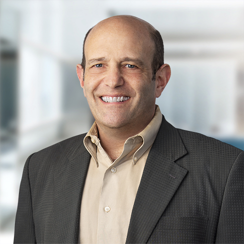 A middle-aged man in a suit jacket and beige shirt smiles at the camera in a modern, blurred corporate law office setting.