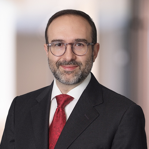 A man with short dark hair, glasses, and a beard, wearing a dark suit, white shirt, and red tie, stands in front of a blurred indoor background typical of modern law offices.