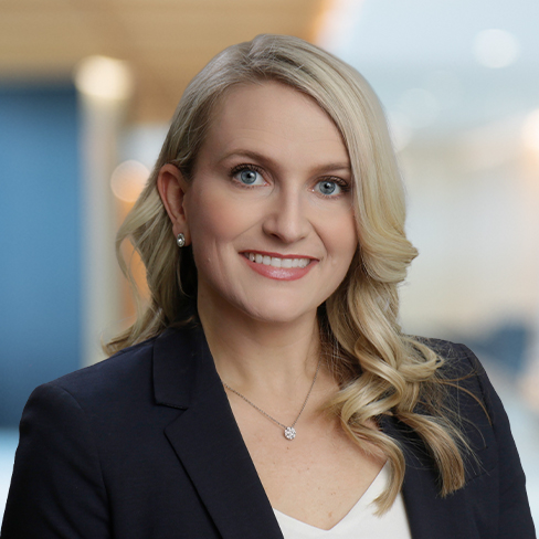 A woman with long blonde hair wearing a dark blazer, white top, and necklace, smiling in a professional indoor setting—ideal for law offices or litigation support environments.