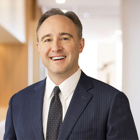 Smiling man wearing a dark pinstripe suit, white shirt, and patterned tie stands in a brightly lit hallway with neutral colors, reflecting the professional atmosphere of a corporate law office.